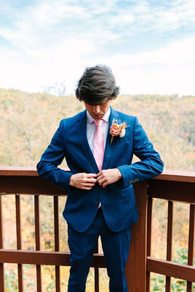 Groom buttoning his blue suit jacket on a mountain-view cabin deck