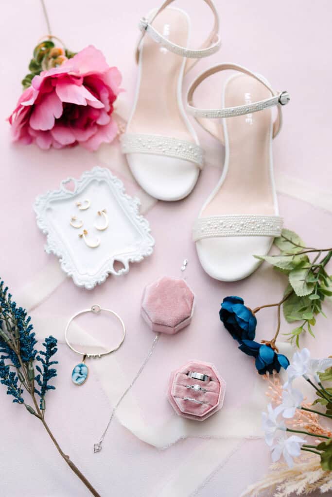 Blush wedding shoes, invitation details, ring boxes, and flowers arranged in a bridal flat lay