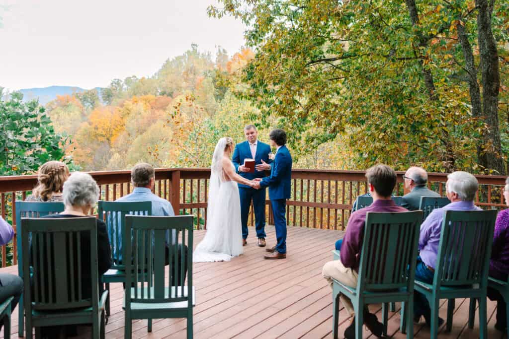 Bride and groom standing with their officiant as guests watch from the deck