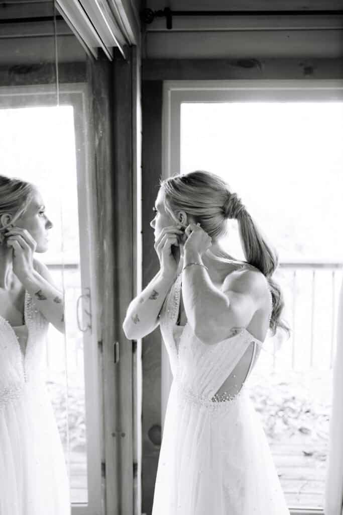 Black and white portrait of the bride putting on an earring beside a window