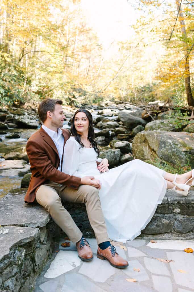 Thaís + Nick Wedding Bride and groom sitting on a stone ledge under golden fall trees